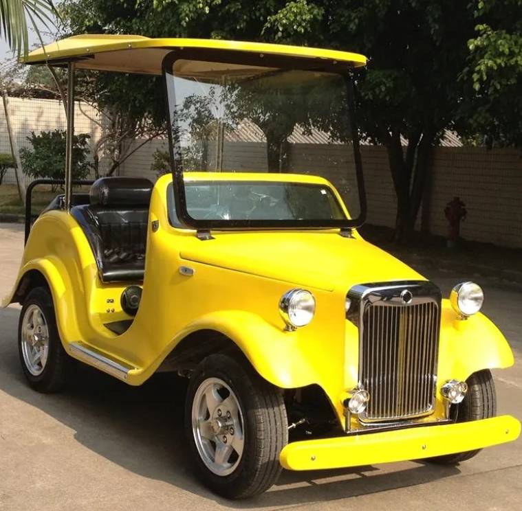 ocean cartz side profile view of a black classic antique-style golf cart with a black roof and large, sweeping fenders. This two-seater vintage electric vehicle features a windshield, light beige or tan upholstery, and black custom wheels with silver accents. The classic golf cart is parked on asphalt outside a modern commercial building with trees and bushes in the foreground.