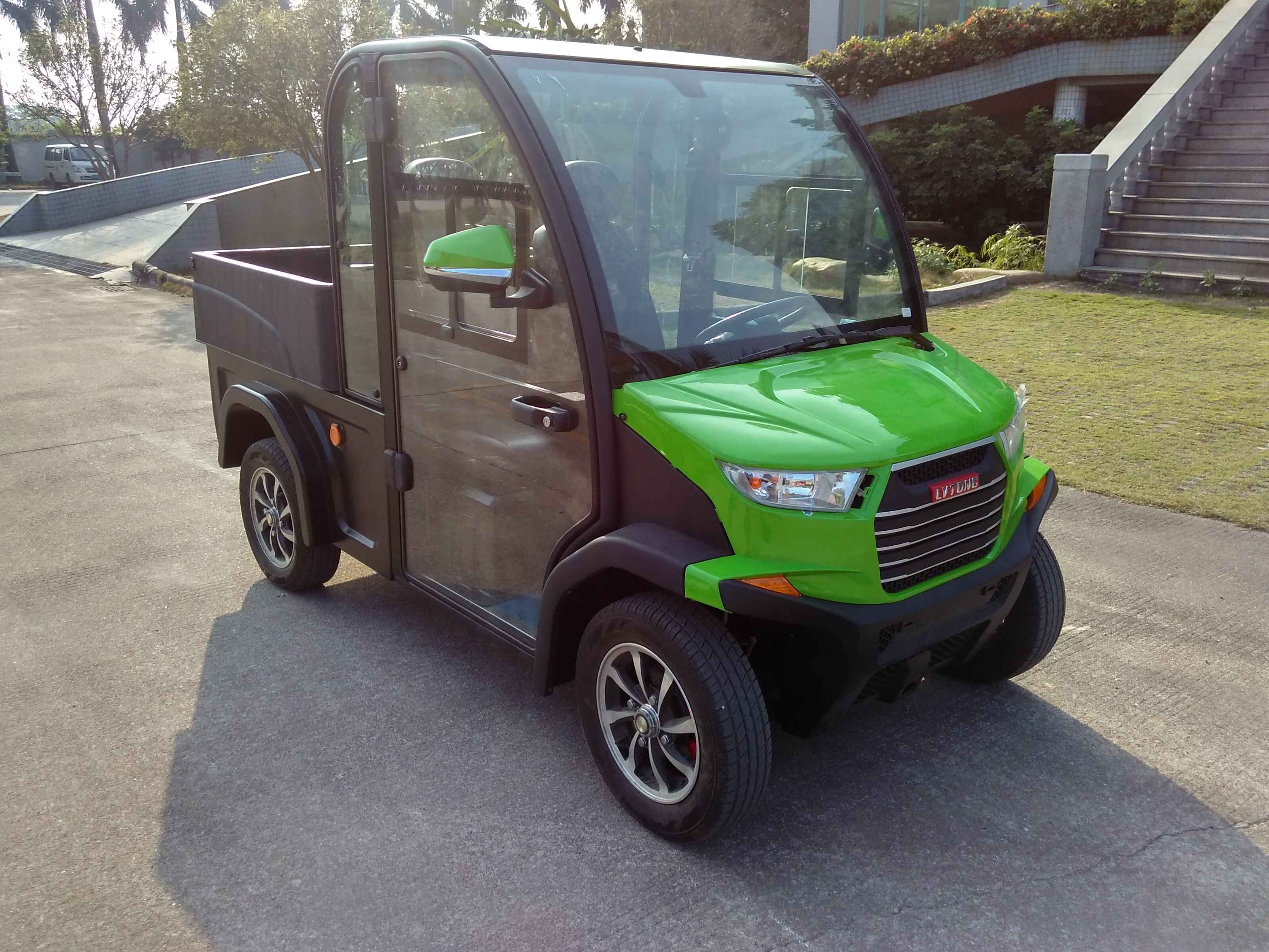 Green and brown utility vehicle on a paved surface with a building and stairs in the background.