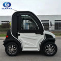 ocean cartz side profile view of a white and black two-seater electric vehicle with a fully enclosed cab, parked on an asphalt road. This compact low-speed electric car or NEV features a large driver's door with a pronounced white panel design, black fender flares, and a small rear window. It is equipped with custom black and chrome alloy wheels, emphasizing its all-weather neighborhood electric car design.