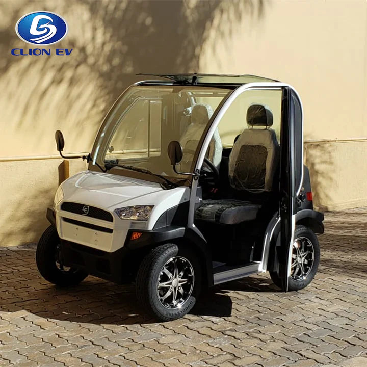 ocean cartz three-quarter front view of a white and black two-seater electric vehicle with a fully enclosed cab and a small, tinted roof. This compact NEV or low-speed electric car has a modern front end, black-trimmed doors with clear glass, and black seats. It is equipped with custom black alloy wheels and is parked on a paved stone driveway in a sunny, residential or resort setting.