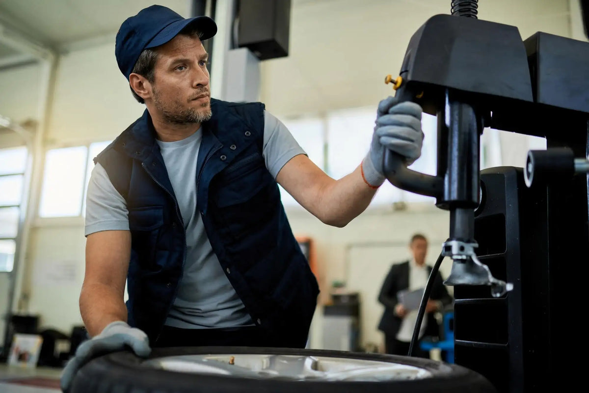 A mechanic in a navy blue cap and vest operates a black tire-changing machine with a silver metallic handle.